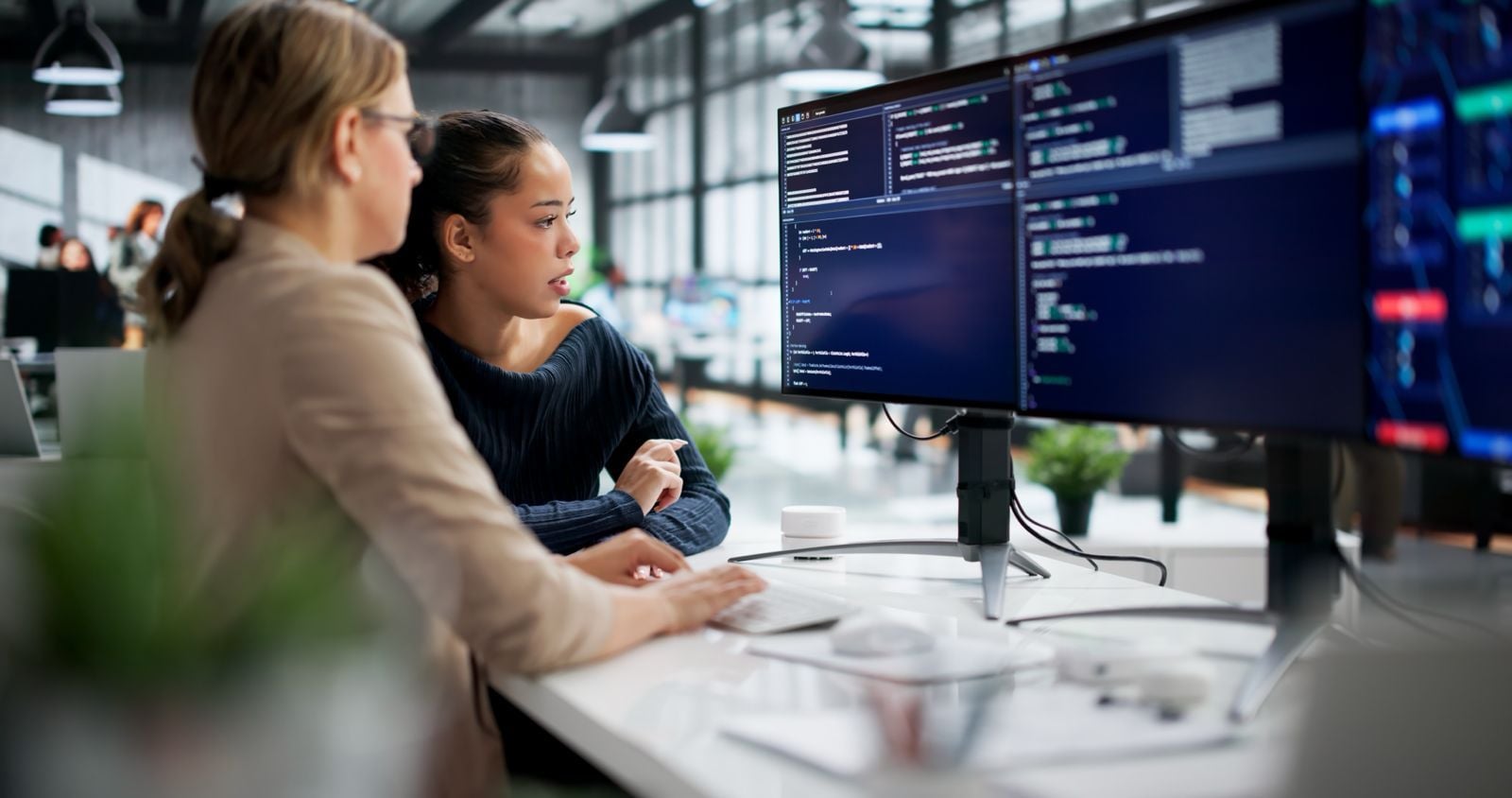 Two female AI engineers collaborating on software development and reviewing code on dual monitors in a tech office