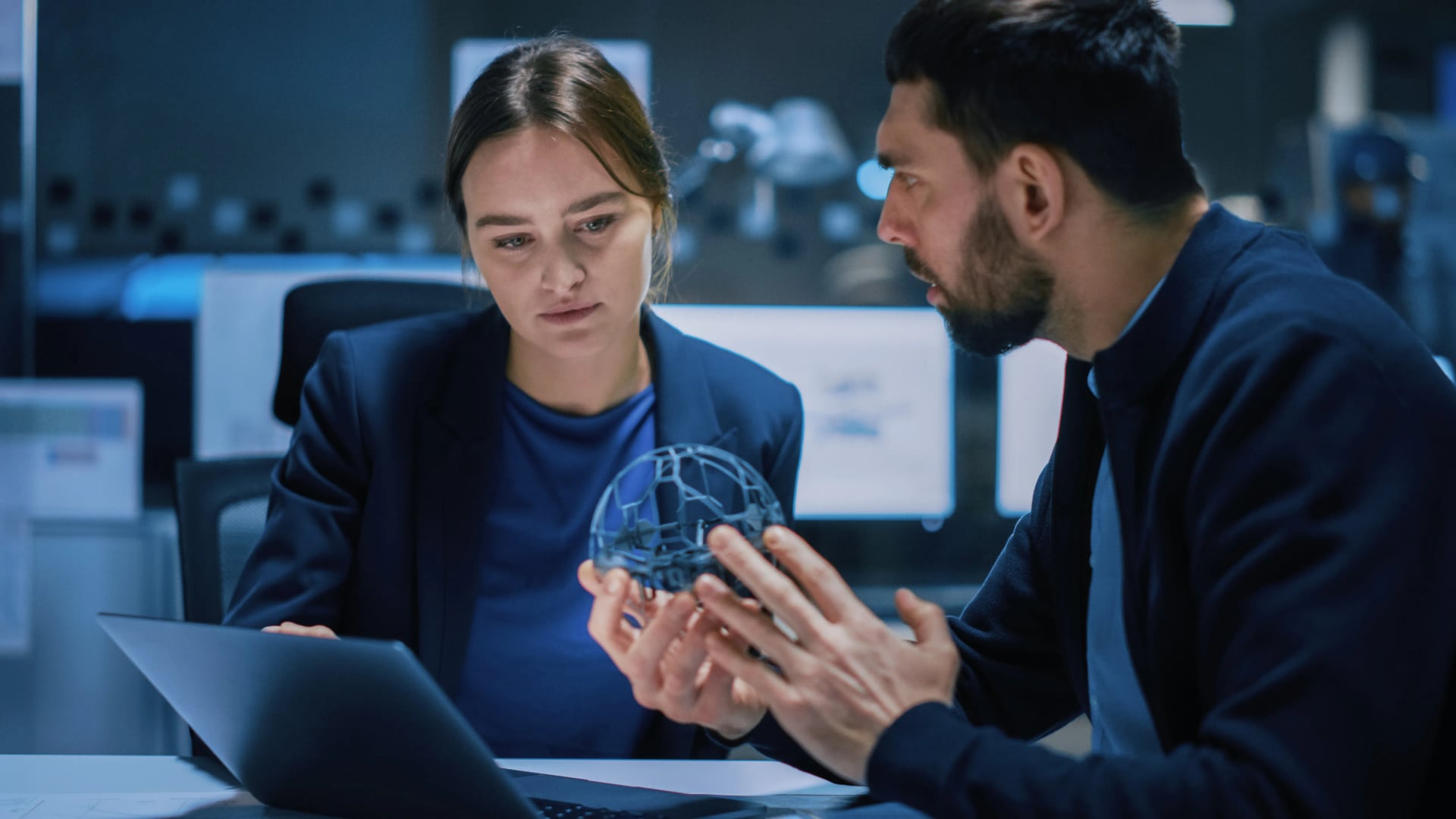 A forward-deployed engineer and a colleague collaborating over a laptop while closely examining a spherical wireframe object in a modern, dimly lit technology office.