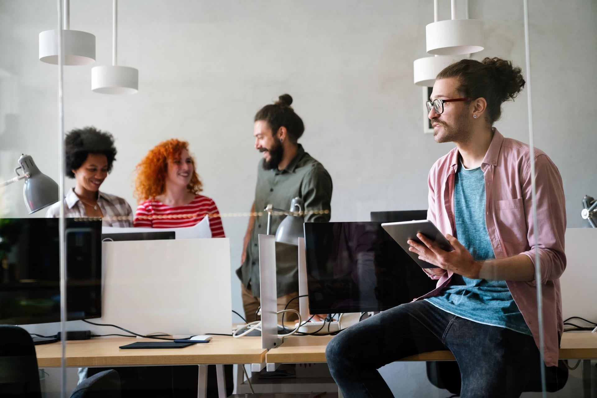 A diverse group of creative professionals collaborating in a modern, open-plan office. A man with glasses and a bun sits on a desk in the foreground holding a tablet, while colleagues chat behind a glass partition.