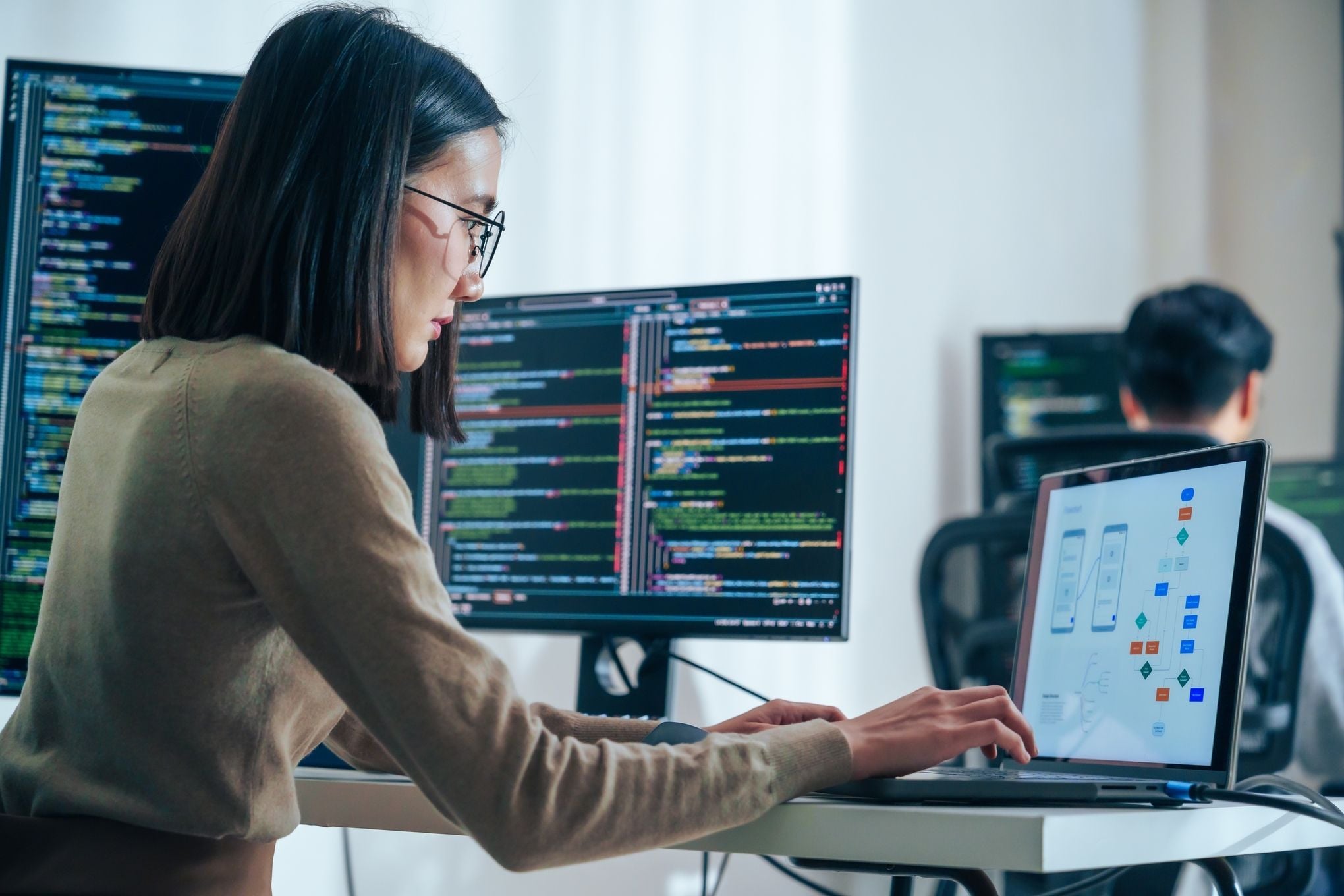 A professional female software engineer working in a modern office, analyzing code on multiple monitors and a workflow diagram on a laptop, representing the high-quality technical talent in Latin American nearshore hubs.